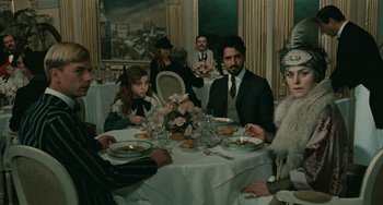 Movie still from “The Ship Sails On” (1983), directed by Federico Fellini – A group of people sitting at a table with plates of food on it; Medium shot, Over the shoulder angle