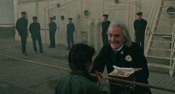 Movie still from “The Ship Sails On” (1983), directed by Federico Fellini – An older man holding a box of food while a young girl looks on; Medium shot, Over the shoulder angle