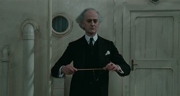 Movie still from “The Ship Sails On” (1983), directed by Federico Fellini – A man in a suit and tie holding a wooden stick; Medium shot, Low angle