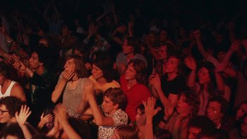 Movie still from “The Song Remains the Same” (1976), directed by Peter Clifton – A group of people sitting in a room with their hands raised; Medium shot, High angle