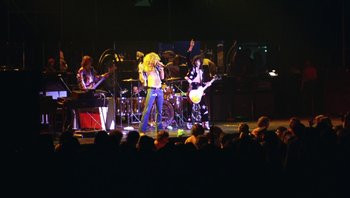 Movie still from “The Song Remains the Same” (1976), directed by Peter Clifton – A group of people on a stage with a band; Wide shot, Low angle