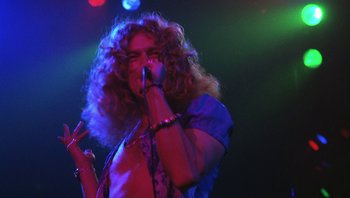 Movie still from “The Song Remains the Same” (1976), directed by Peter Clifton – A man with long curly hair is holding a microphone; Medium shot, Low angle