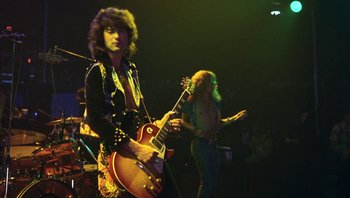 Movie still from “The Song Remains the Same” (1976), directed by Peter Clifton – A man in a black jacket playing an electric guitar on a stage; Medium shot, Low angle