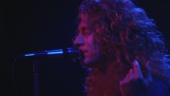 Movie still from “The Song Remains the Same” (1976), directed by Peter Clifton – A man with long curly hair holding a microphone in front of him; Close Up shot, Low angle