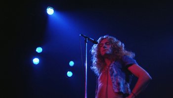 Movie still from “The Song Remains the Same” (1976), directed by Peter Clifton – A man with long curly hair standing on a stage with a microphone; Medium shot, Low angle