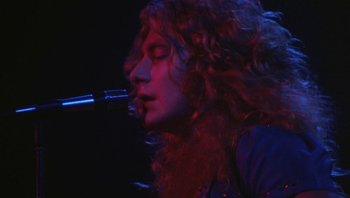 Movie still from “The Song Remains the Same” (1976), directed by Peter Clifton – A person with long hair holding a microphone; Close Up shot, Low angle