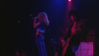 Movie still from “The Song Remains the Same” (1976), directed by Peter Clifton – A woman standing on a stage singing into a microphone with a man standing next to her; Wide shot, Low angle