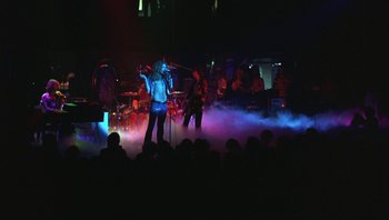 Movie still from “The Song Remains the Same” (1976), directed by Peter Clifton – A group of people on a stage with a microphone; Wide shot, High angle