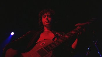 Movie still from “The Song Remains the Same” (1976), directed by Peter Clifton – A man holding a guitar while standing in a dark room; Medium shot, Low angle