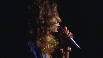 Movie still from “The Song Remains the Same” (1976), directed by Peter Clifton – A woman with curly hair is singing into a microphone; Close Up shot, Low angle