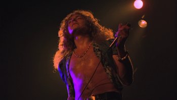 Movie still from “The Song Remains the Same” (1976), directed by Peter Clifton – A man with long curly hair is holding a microphone; Medium shot, Low angle