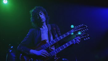 Movie still from “The Song Remains the Same” (1976), directed by Peter Clifton – A man holding a guitar while standing on a stage; Medium shot, Low angle