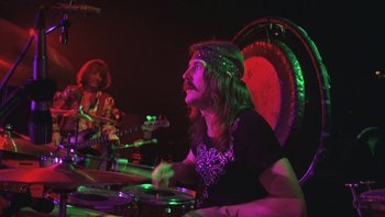 Movie still from “The Song Remains the Same” (1976), directed by Peter Clifton – A man sitting at a table playing drums; Medium shot, Low angle