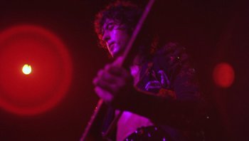 Movie still from “The Song Remains the Same” (1976), directed by Peter Clifton – A man holding a guitar in front of a red light; Medium shot, Low angle