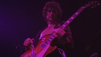 Movie still from “The Song Remains the Same” (1976), directed by Peter Clifton – A person with a guitar in a dark room; Close Up shot, Low angle