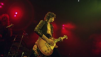 Movie still from “The Song Remains the Same” (1976), directed by Peter Clifton – A man in a black jacket is playing a guitar; Medium shot, Low angle