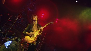 Movie still from “The Song Remains the Same” (1976), directed by Peter Clifton – A man playing a guitar on a stage; Medium shot, Low angle
