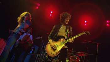 Movie still from “The Song Remains the Same” (1976), directed by Peter Clifton – A man playing a guitar on a stage with a band; Medium shot, Low angle