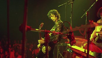 Movie still from “The Song Remains the Same” (1976), directed by Peter Clifton – A man playing a guitar on stage in front of an audience; Medium shot, Low angle