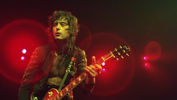 Movie still from “The Song Remains the Same” (1976), directed by Peter Clifton – A man playing a guitar in front of a red light; Medium shot, Low angle