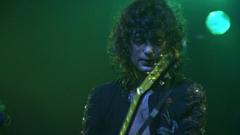 Movie still from “The Song Remains the Same” (1976), directed by Peter Clifton – A man with curly black hair is holding a guitar; Close Up shot, Low angle