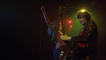 Movie still from “The Song Remains the Same” (1976), directed by Peter Clifton – A man and woman playing guitar in a band; Medium shot, Low angle