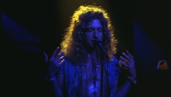 Movie still from “The Song Remains the Same” (1976), directed by Peter Clifton – A man with curly hair is singing into a microphone; Medium shot, Low angle