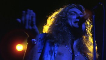 Movie still from “The Song Remains the Same” (1976), directed by Peter Clifton – A man with long curly hair singing into a microphone; Close Up shot, Low angle