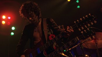 Movie still from “The Song Remains the Same” (1976), directed by Peter Clifton – A man playing a guitar in a dark room; Medium shot, Low angle