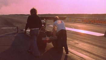 Movie still from “The Song Remains the Same” (1976), directed by Peter Clifton – Two people working on a race car on a race track; Wide shot, Low angle