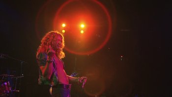 Movie still from “The Song Remains the Same” (1976), directed by Peter Clifton – A man with long curly hair is holding a microphone; Medium shot, Low angle