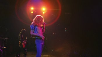 Movie still from “The Song Remains the Same” (1976), directed by Peter Clifton – A man with a microphone on a stage with lights in the background; Wide shot, Low angle