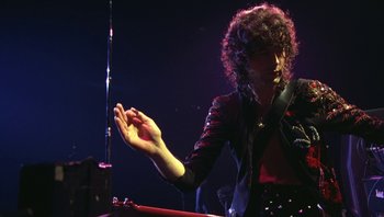 Movie still from “The Song Remains the Same” (1976), directed by Peter Clifton – A man with curly hair is playing the drums; Medium shot, Low angle