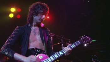 Movie still from “The Song Remains the Same” (1976), directed by Peter Clifton – A man playing a guitar in front of a crowd; Medium shot, Low angle