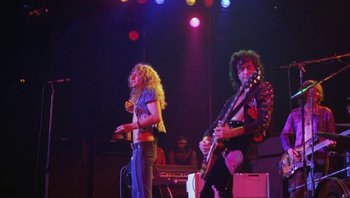 Movie still from “The Song Remains the Same” (1976), directed by Peter Clifton – A man and a woman on a stage with a guitar; Medium shot, Low angle
