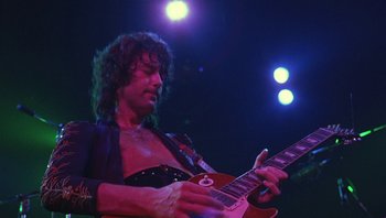 Movie still from “The Song Remains the Same” (1976), directed by Peter Clifton – A man playing a guitar on a stage with lights in the background; Medium shot, Low angle