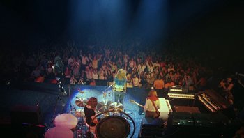 Movie still from “The Song Remains the Same” (1976), directed by Peter Clifton – A group of people on a stage playing drums and singing; Wide shot, High angle