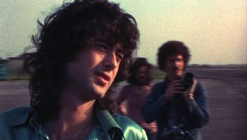 Movie still from “The Song Remains the Same” (1976), directed by Peter Clifton – A man with long curly hair is looking at the camera; Close Up shot, Over the shoulder angle