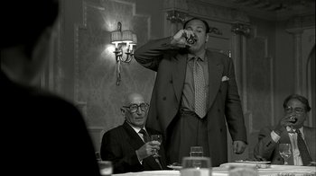 Movie still from “The Sopranos” (1999), created by David Chase – A man in a suit and tie is drinking a glass of wine while another man in a suit and tie looks on; Medium shot, Low angle