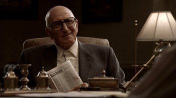 Movie still from “The Sopranos” (1999), created by David Chase – An older man sitting at a desk holding a newspaper; Medium shot, Over the shoulder angle
