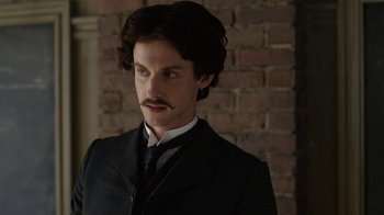 Movie still from “Anne with an E” (2017), created by Moira Walley-Beckett – A man in a suit and tie standing in front of a brick wall; Close Up shot, Low angle