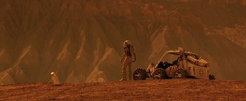 Movie still from “The Space Between Us” (2017), directed by Peter Chelsom – A person standing on a dirt hill next to a vehicle; Wide shot, High angle