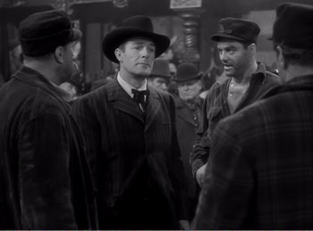 Movie still from “The Spoilers” (1942), directed by Ray Enright – A black and white photo of a group of men in hats; Medium shot, Over the shoulder angle