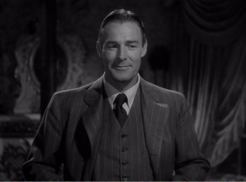 Movie still from “The Spoilers” (1942), directed by Ray Enright – A man wearing a suit and tie in front of a curtain; Medium shot, Low angle