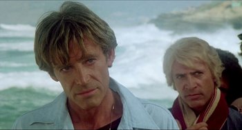 Movie still from “The Stunt Man” (1980), directed by Richard Rush – A man standing next to another man on a beach; Close Up shot, Low angle