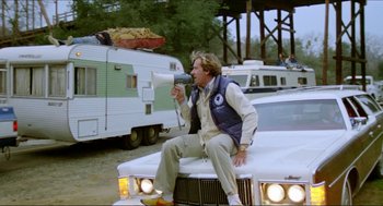 Movie still from “The Stunt Man” (1980), directed by Richard Rush – A man sitting on the hood of a car holding a megaphone; Wide shot, Low angle