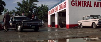 Movie still from “The Sugarland Express” (1974), directed by Steven Spielberg – A man sitting in the back of a car in front of a garage; Wide shot, Low angle