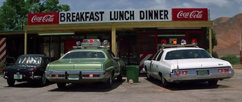 Movie still from “The Sugarland Express” (1974), directed by Steven Spielberg – Two cars parked in front of a diner with a dog laying in front of them; Wide shot, Low angle