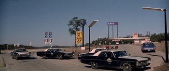 Movie still from “The Sugarland Express” (1974), directed by Steven Spielberg – A couple of police cars parked on the side of a road; Wide shot, Over the shoulder angle
