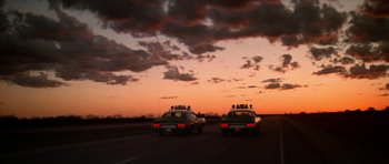 Movie still from “The Sugarland Express” (1974), directed by Steven Spielberg – A couple of cars driving down a street under a cloudy sky; Extreme Wide shot, Low angle